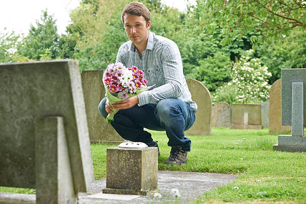 istock Man Placing Flowers By Headstone In Cemetery 181068727