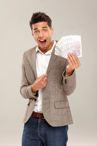 Surprised young businessman pointing at money fanned in his hand against grey background