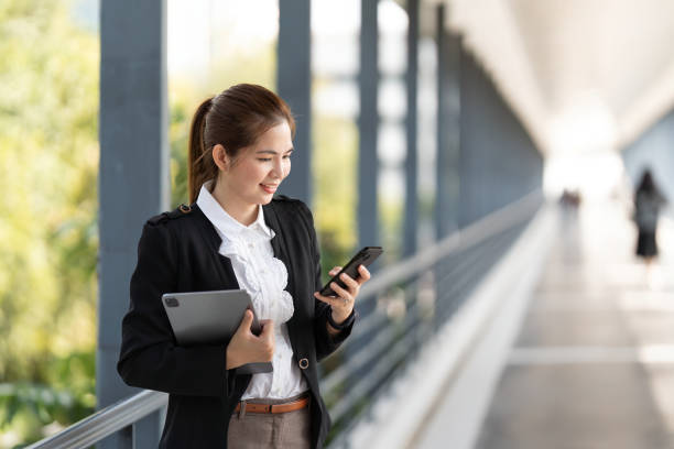 young asian business woman wearing suit using application on cell phone, read news on smartphone, fast connection, checking mobile apps outdoors - news of the world imagens e fotografias de stock