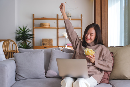 Portrait image of a happy woman eating potato chips while working or watching movie on laptop at home