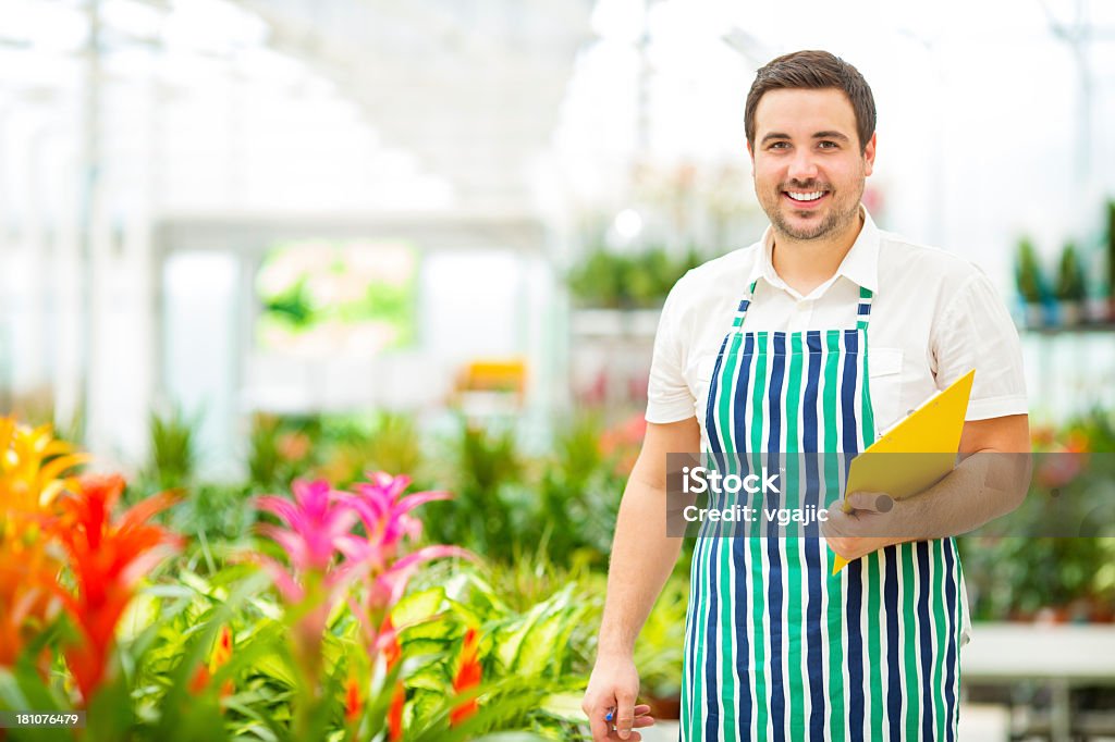 Male Florist Working At Garden Center. Portrait of an young male florist nursery colourful flowers. Florist standing behind colourful orchids, holding notepad and writing what he needs to order. 25-29 Years Stock Photo