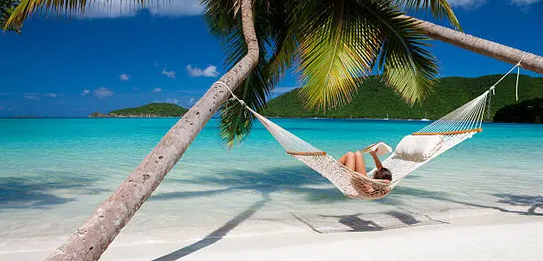 woman reading a book in a hammock stretched between two palm trees at a tropical beach in the Caribbean