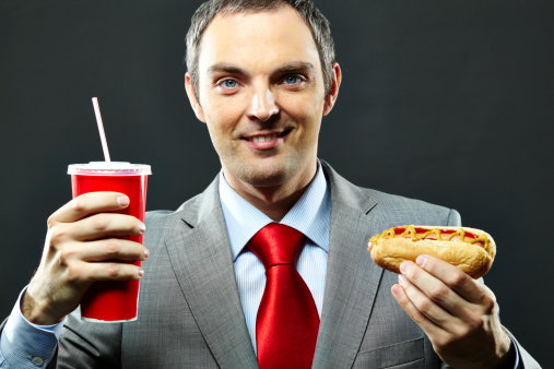 Portrait of smiling businessman eating unhealthy food: soda and hot dog