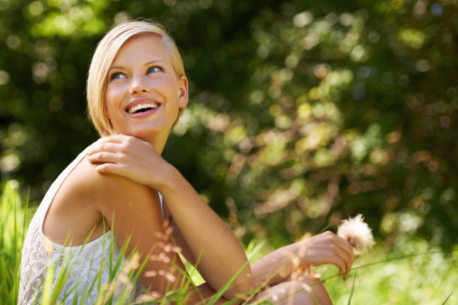 An attractive young woman sitting in an open field and holding a dandelion