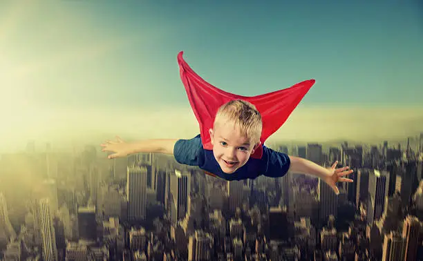 Boy looks happy and a bit surprised as he flys over a big city with tall buildings. He is wearing a super hero outfit and a red cape.