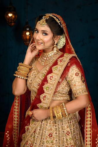 Stunning Indian bride dressed in traditional bridal lehenga with heavy gold jewellery and veil smiles tenderly in studio lighting. Wedding fashion and lifestyle.