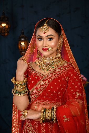 Stunning Indian bride dressed in traditional red bridal lehenga with heavy gold jewellery and veil smiles tenderly in studio lighting. Wedding fashion and lifestyle.