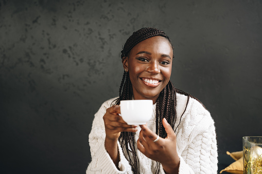 African woman in warm white sweater drinking hot coffee or tea at home close up