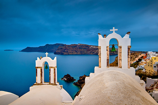 Two bell towers and dome roofs of white Greek Orthodox church in Oia village, Santorini island, against waters of Aegean sea, Greece, and distant island on overcast day with heavy clouds and dramatic sky. Famous local landmark, wedding destination