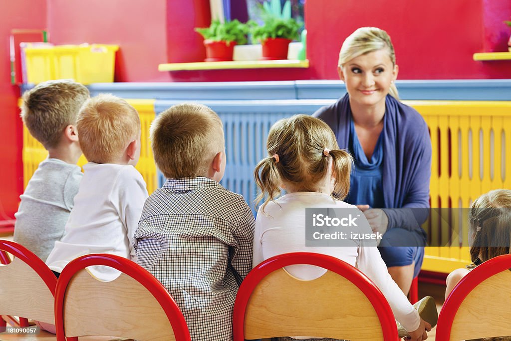 Nursery School A nursery school children sitting on chairs in a playroom and listening to their teacher. 20-24 Years Stock Photo