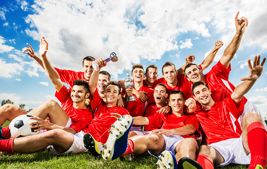 Cheerful soccer team sitting on grass and holding winning trophies.