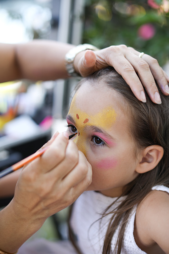 Adorable girl getting her face painted. Child animator, artist's hand draws face painting to little girl. Child with funny face painting. Painter makes blue butterfly at girl's face. Children holiday, event, birthday party, entertainment.
