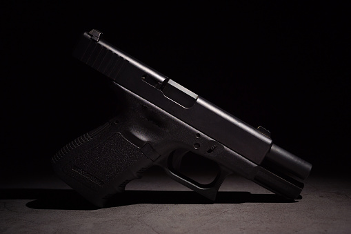 Close-up of a black metal 9mm automatic pistol with bullets all placed on a cement table with a black background.