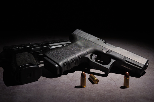 Close-up of two black metal 9mm automatic pistols with bullets all placed on a cement table with a black background.
