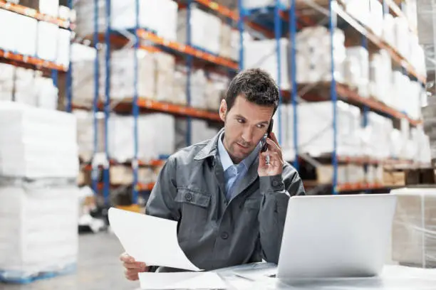 A young man in a warehouse taking orders on his cellphone and recording them on his laptop