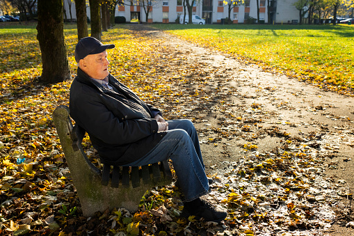 87 years old adult senior with hearing aid in his ear sitting on bench enjoying warm autumn afternoon in park.