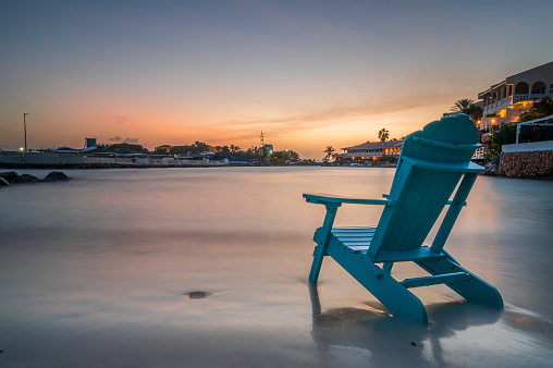 An Adirondack chair, on the shoreline of a tropical beach, at sunset. The ocean is lapping at the chair.