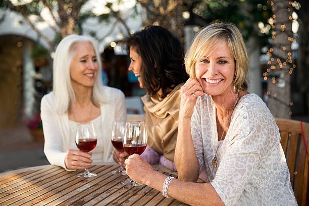 Girlfriends drinking wine Three Beautiful, mature women drinking wine while taking at a table in a cafe outdoors. Its their girls night out. Women are chatting, laughing and having a great time. One of the woman is in main focus and looking at the camera with a warm smile. Women are between 50 and 65 years old. women only restaurant stock pictures, royalty-free photos & images