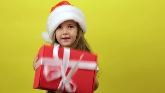 Portrait cute little girl kid in Santa hat holding gift box posing isolated over yellow background wall in studio. Christmas concept