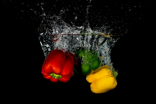 Whole red, green and yellow bell peppers falling into water with a black background creating bubbles and a splash