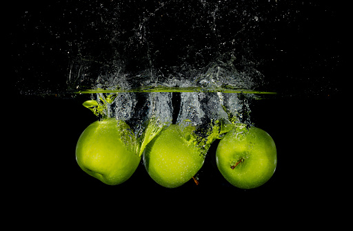 Three whole green apples falling into water with a black background creating a splash and bubbles
