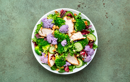 Healthy fresh salad from purple cauliflower, broccoli, grilled chicken breast, cucumbers, red onion, radicchio, spinach with walnuts, green table background, top view