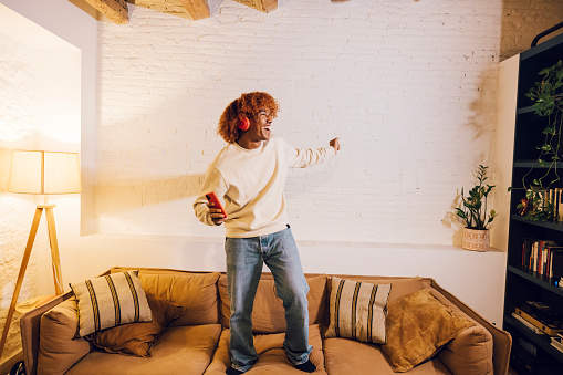 Young african american man listening music with red headphones and dancing on a sofa in living room. Very effusive man singing and dancing at home.