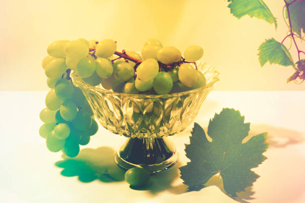 A bunch of white grapes in a glass bowl with a silver base. A grape vine and leaf serve as decoration