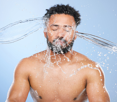 Black man, water splash and washing in skincare for cleaning, hygiene or grooming against a blue studio background. Face of African male person, model and aqua for body hydration, shower or skin wash