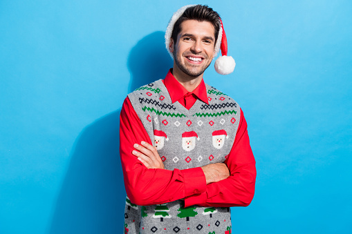 Photo portrait of handsome young guy crossed arms posing shopping wear x-mas print vest hat outfit isolated on blue color background.