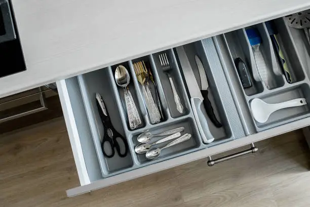 Open drawer in a kitchen, which is part of a white kitchen cabinet and sitting on a wooden floor. Inside the drawer, a variety of kitchen utensils, including forks, spoons, and knives