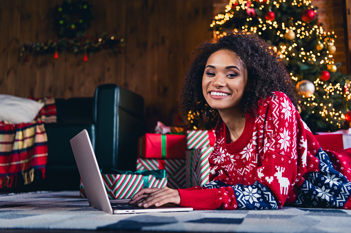 Photo of charming black skin girl lying on fluffy carpet chatting with friends on macbook xmas day magic spirit decorations room indoors