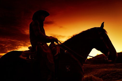 Cowboy Sitting On His Horse In A Flamming, Hot,Western Sunset - Silhouette.  Copy Space.  Added grain..Brand has been altered from its original look, the Rocking K, brand.  It is now the Sacred Ground K brand.  (This brand is actually non-existant as displayed on this image.)
