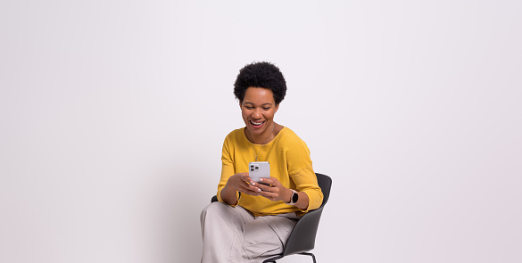 Cheerful businesswoman checking messages over smart phone and sitting on chair over white background