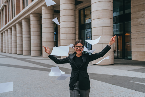 Happy brunette young entrepreneur throwing up paper sheets looks at camera happily rising hands up celebrating success. Cheerful American female student glad  by passed exams.