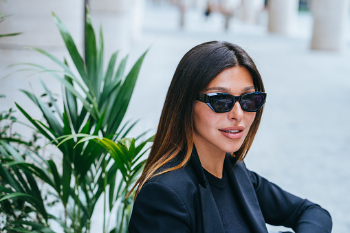 Close up portrait of stylish luxury dressed female model in sunglasses sitting outside against green plants. Tanned successful woman relaxing at outside cafe. Wealthy lifestyle.