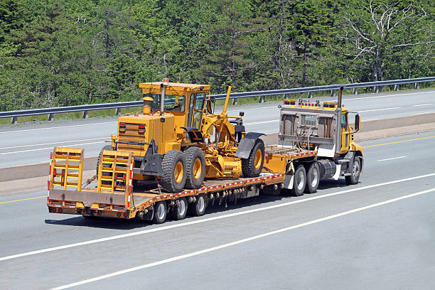 Transporting A Road Grader Elevated rear quarter view of a flatbed semi truck on a highway transporting a road grader. large machine stock pictures, royalty-free photos & images