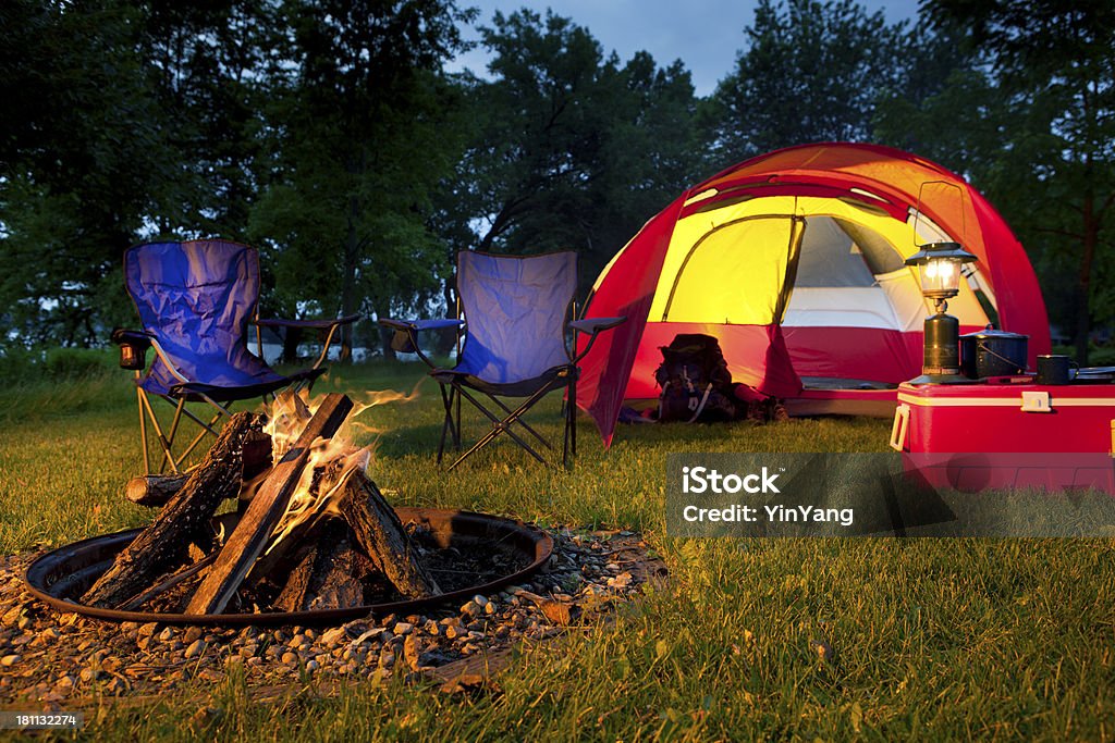 Evening Campsite with Red Tent, Chairs, and Burning Fire Pit Subject: Horizontal view of evening campsite. A red tent set up on green grass, a fire pit burning a campfire in the foreground, chairs, cooler, lamp and cooking equipment suggest relaxation in a natural night setting. Camping Stock Photo