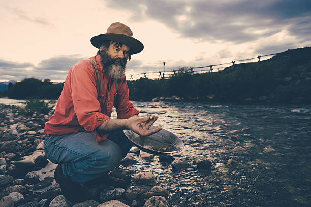 Gold Panning,Successful Prospector with Nugget "Senior Prospector or Gold Miner, panning for Gold at a lonely river during sunset. Holding a gold nugget between his fingers. Old-fashioned image style with desaturated colors." panning for gold photos stock pictures, royalty-free photos & images