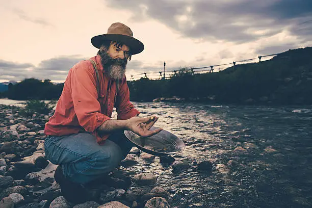 "Senior Prospector or Gold Miner, panning for Gold at a lonely river during sunset. Holding a gold nugget between his fingers. Old-fashioned image style with desaturated colors."
