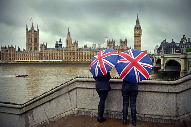 London rain "Tourists huddle beneath British flag umbrellas (they sell them there) during a London summer rainstorm near the Houses of Parliament. A dark cloudy typical summer day!Westminster, LondonUK" union jack flag stock pictures, royalty-free photos & images