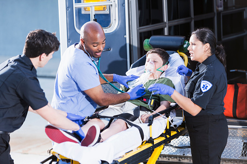 Multi-ethnic doctor and paramedics (30s, 40s) helping boy (12 years) lying on a stretcher outside an ambulance.