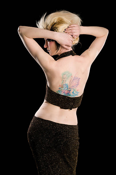 Rear view of model in black and gold halter dress. Vertical studio shot on black of blonde woman with colourful tattoo and hands in hair. black pin up girl tattoos stock pictures, royalty-free photos & images
