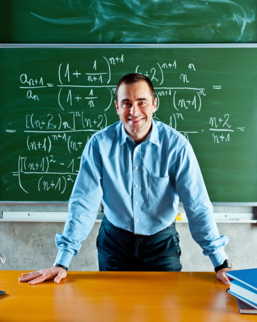 Math teacher standing against blackboard full of formulas and smiling at the camera.