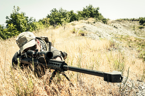 A marksman on burnt grass, is taking aim with his scope mounted on a precision cal .50 rifle.