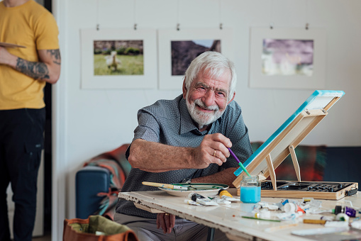 Front view of a senior man attending an art class at an art studio in a home in Horsely, Northumberland, England. He is holding a paintbrush while painting on a canvas to create art. He is smiling and looking at the camera.