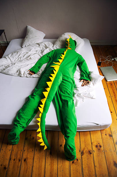 Dinosaur Costume Man Lies Face Down in Bed Dinosaur man lies face down in a simple bed on the floor, gadgets charging beside him face down stock pictures, royalty-free photos & images