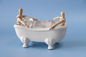 Close-up image of two wooden lay figures, jointed artist's models sitting in model, Victorian style, white ceramic freestanding curved roll top bath with claw feet filled with cubes of ice, light blue background, health and therapy concept