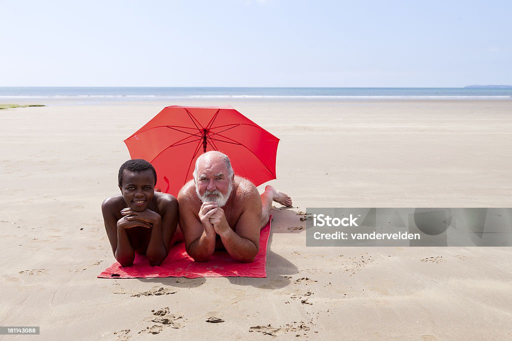 The Odd Couple An older Caucasian man and an African woman relax together on a beach. Nudist Stock Photo