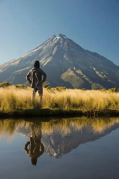 A man looks towards Mt Taranaki, with their reflections visible in a mountain tarn.